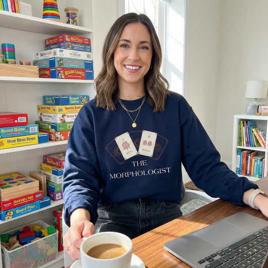 Speech therapist wearing a navy “The Morphologist” sweatshirt sitting at a desk with books and coffee
